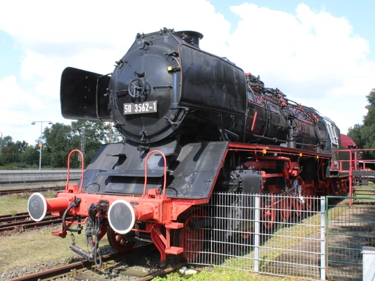 Eine historische Dampflok steht majestätisch auf Gleisen, eingerahmt von einem grünen Zaun.A historic steam locomotive stands majestically on the tracks, framed by a green fence.Et historisk damplokomotiv står majestætisk på skinnerne, indrammet af et grønt hegn.Een historische stoomlocomotief staat majestueus op het spoor, omringd door een groen hek.
