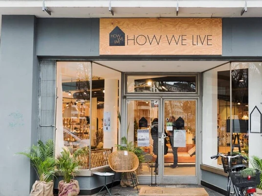 How we live Das Bild zeigt die einladende Schaufensterfassade des Geschäfts „HOW WE LIVE“ mit dezentem grünen Pflanzenarrangement und einem Fahrrad im Vordergrund.The picture shows the inviting store front of the "HOW WE LIVE" store with a subtle green plant arrangement and a bicycle in the foreground.