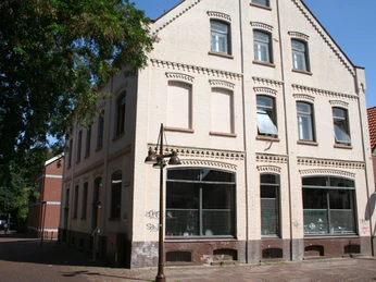 Dreigeschossiges Backsteingebäude mit Ornamentfenstern, liegt an einer gepflasterten Straßenecke.