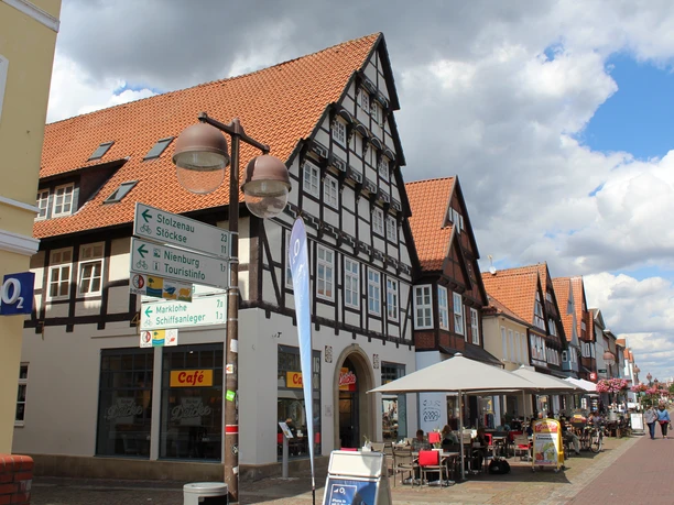Fachwerkhaus mit rotem Dach in Nienburg, umgeben von Geschäften und Straßencafés bei bewölktem Himmel.
