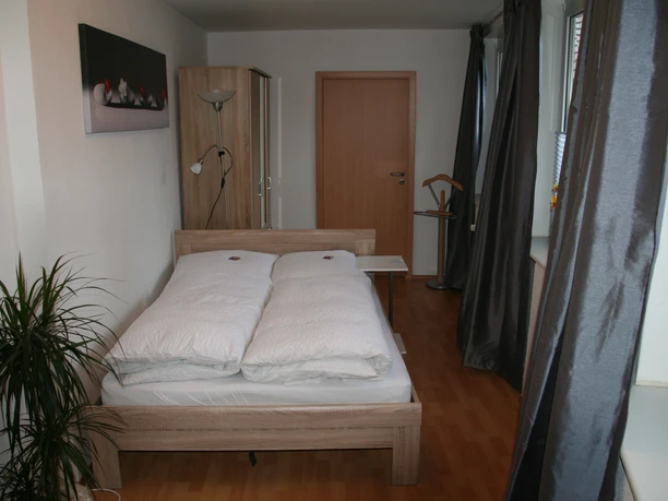 A bright bedroom with a double bed, a closet and a window with dark curtains.