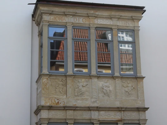 Ein Renaissance-Erker aus Sandstein mit Verzierungen und Inschriften an einer weißen Hausfassade.