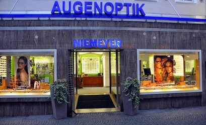 Augenoptik Niemeyer Das Bild zeigt die Außenansicht des Optikersgeschäfts Augenoptik Niemeyer mit einer beleuchteten Glasfassade und einem modernen Design. In den Schaufenstern sind Werbebilder und Brillenmodelle zu sehen, während über dem Eingang ein großes, blaues Schild mit der Aufschrift "AUGENOPTIK NIEMEYER" leuchtet.