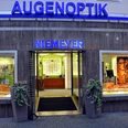 Augenoptik Niemeyer Das Bild zeigt die Außenansicht des Optikersgeschäfts Augenoptik Niemeyer mit einer beleuchteten Glasfassade und einem modernen Design. In den Schaufenstern sind Werbebilder und Brillenmodelle zu sehen, während über dem Eingang ein großes, blaues Schild mit der Aufschrift "AUGENOPTIK NIEMEYER" leuchtet.