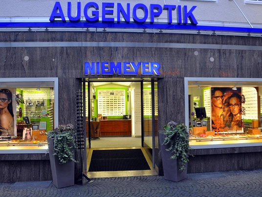 Augenoptik Niemeyer Das Bild zeigt die Außenansicht des Optikersgeschäfts Augenoptik Niemeyer mit einer beleuchteten Glasfassade und einem modernen Design. In den Schaufenstern sind Werbebilder und Brillenmodelle zu sehen, während über dem Eingang ein großes, blaues Schild mit der Aufschrift "AUGENOPTIK NIEMEYER" leuchtet.