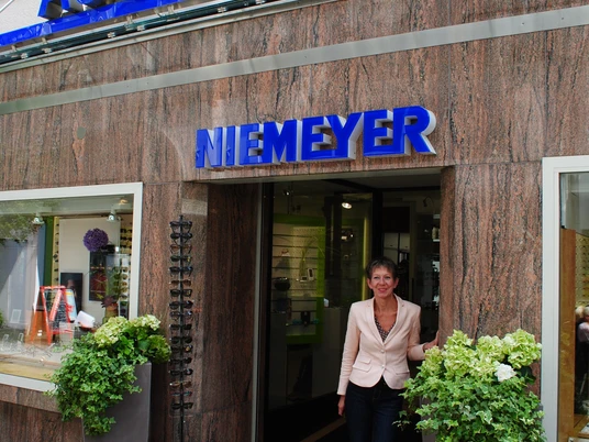 Brillenmode in der Klosterstraße bei Augenoptik Niemeyer Zu sehen ist die Fassade des Optikergeschäfts Niemeyer mit einem blauen Leichtschild über dem Eingang. Vor dem Laden steht eine Frau in heller Kleidung und lächelt in die Kamera.