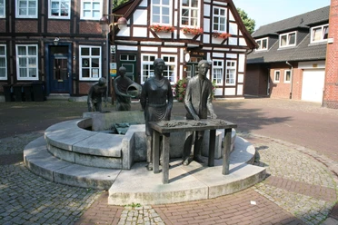 Sculpture of an asparagus farmer with a table and two other figures in front of historic half-timbered houses.