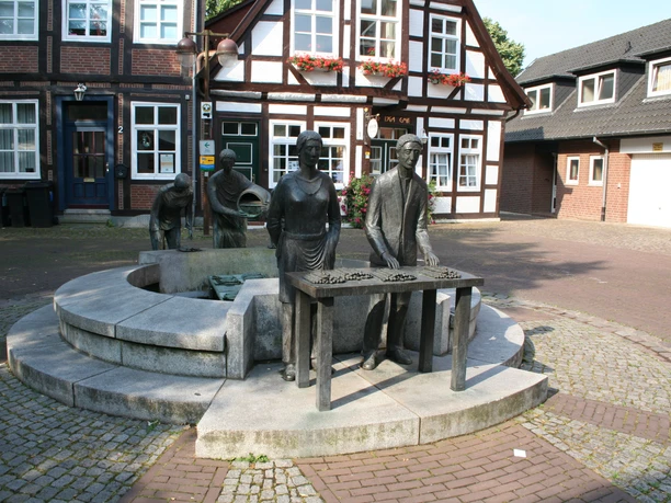Spargelbrunnen Skulptur eines Spargelbauern mit Tafel und zwei weiteren Figuren vor historischen Fachwerkhäusern.