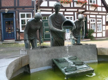 Drei Bronzefiguren schütten Spargel auf einen Gitterrost in einem halbrunden Brunnenensemble aus.Three bronze figures pour asparagus onto a grid in a semi-circular fountain ensemble.Tre bronzefigurer hælder asparges ud på en rist i et halvcirkelformet fontæneensemble.Drie bronzen figuren gieten asperges op een rooster in een halfrond fonteinensemble.