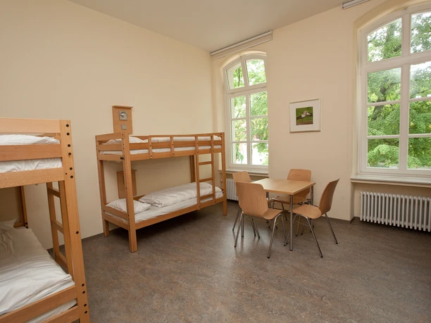 Jugendgästehaus Petershagen Ein heller Raum mit Holzetagenbetten, Tisch und Stühlen vor großen Fenstern mit Blick ins Grüne.