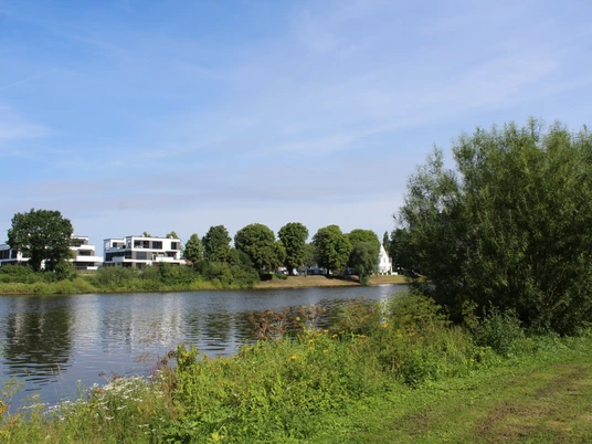 Weser Flusslauf der Weser mit grünen Ufern, modernen Gebäuden und einem Kirchturm im Hintergrund.