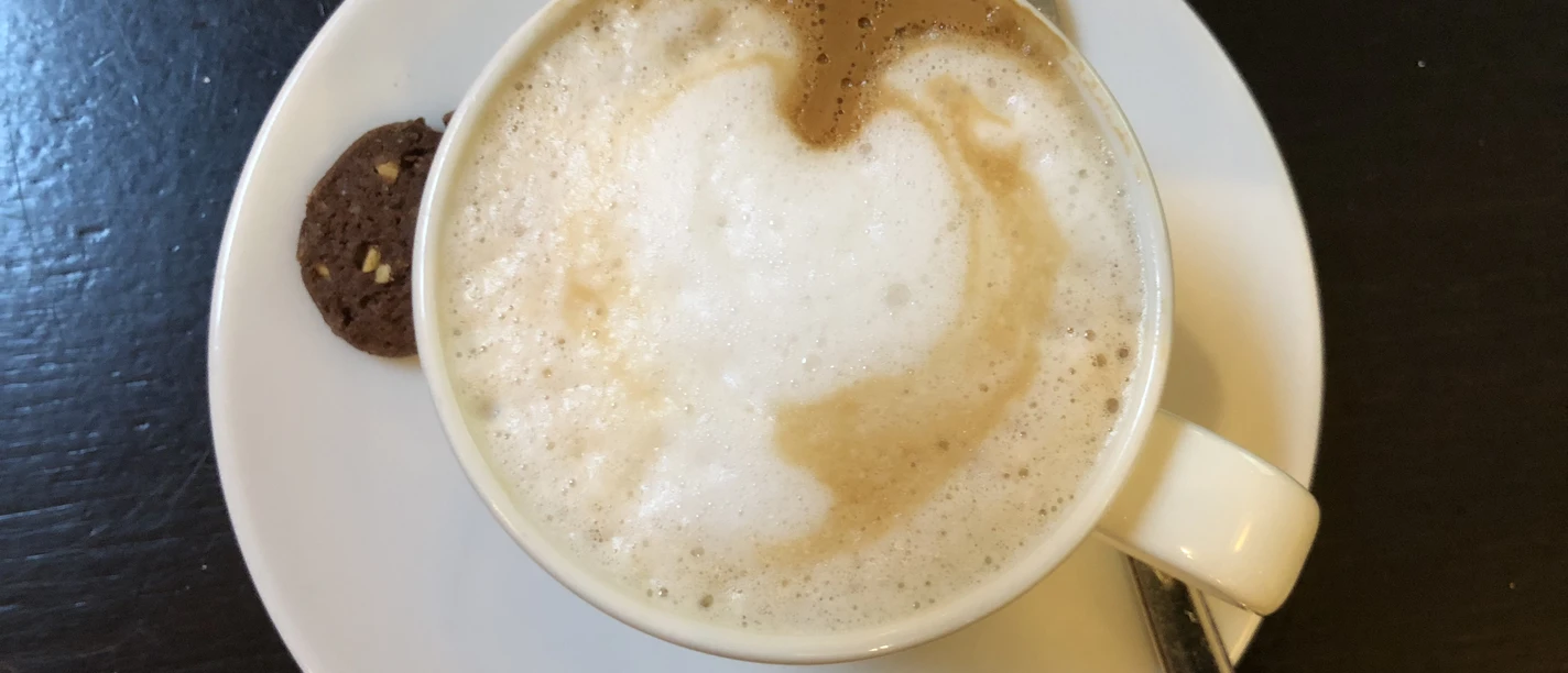 Coffee cup with foam, next to it a chocolate cookie and a spoon on a white saucer.
