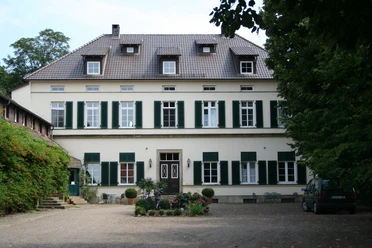 Rittergut Schlüsselburg Herrenhaus mit zwei Stockwerken, grünen Fensterläden, Kiesweg und Garten, umgeben von Bäumen.