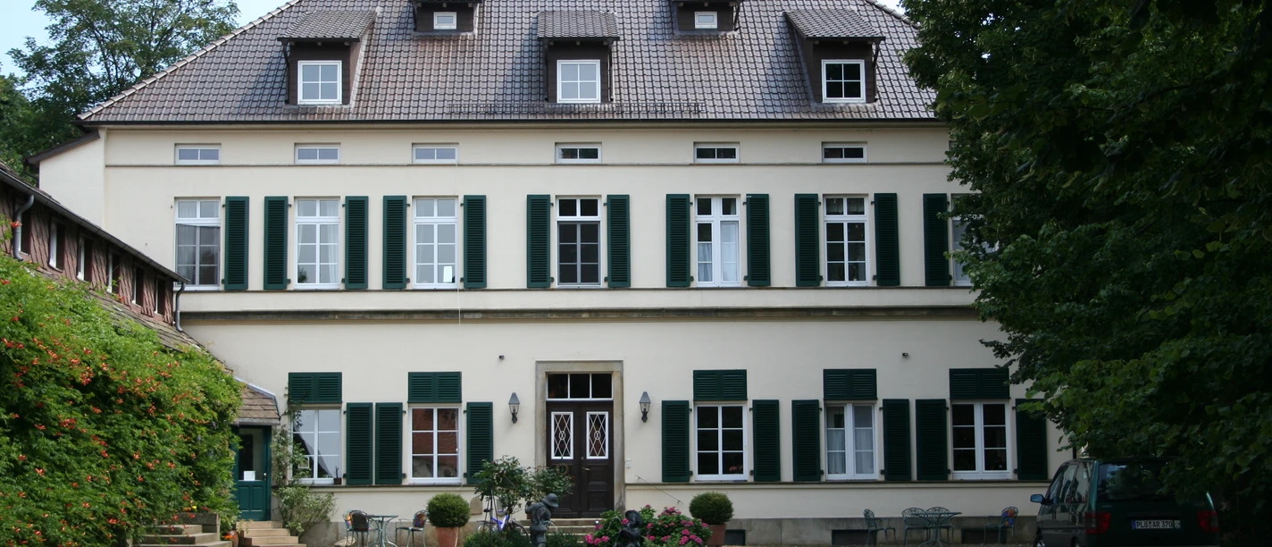 Rittergut Schlüsselburg Herrenhaus mit zwei Stockwerken, grünen Fensterläden, Kiesweg und Garten, umgeben von Bäumen.