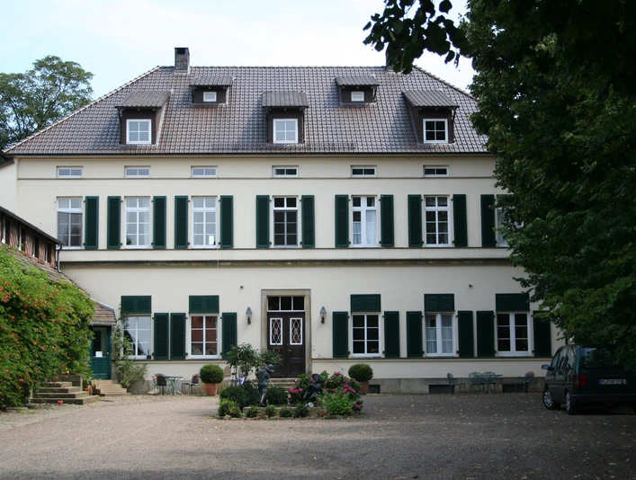 Rittergut Schlüsselburg Herrenhaus mit zwei Stockwerken, grünen Fensterläden, Kiesweg und Garten, umgeben von Bäumen.