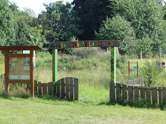 Eingangstor zur Kinderwildnis Nienburg mit üppigem Grün und hölzernem Infobrett im Vordergrund.Entrance gate to the Nienburg Children's Wilderness with lush greenery and wooden information board in the foreground.Indgangsporten til Nienburg Children's Wilderness med frodigt grønt og en informationstavle af træ i forgrunden.Toegangspoort tot de Kinderwildernis Nienburg met weelderig groen en houten informatiebord op de voorgrond.