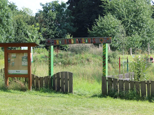 Entrance gate to the Nienburg Children's Wilderness with lush greenery and wooden information board in the foreground.