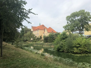Rotes Ziegeldachgebäude hinter grünem Graben mit Seerosen, umgeben von Bäumen und Wiesen.