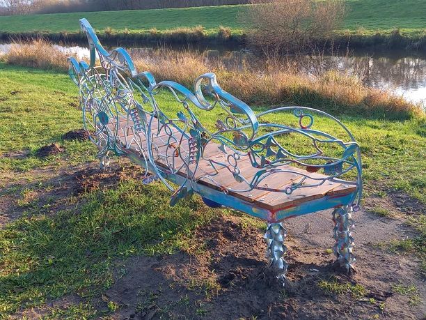 Fischbank am Lieblingsort Heeren Wehren Brücke Die kunstvolle Fischbank an der Heeren Wehren Brücke zeichnet sich durch ihr einzigartiges Design mit geschwungenen, fischförmigen Metallverzierungen aus, die an einen ruhigen Fluss eingebettet sind.