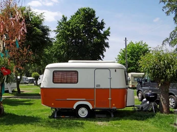 Camping Berger Ein farbenfroher Wohnwagen steht auf einem grünen Rasen zwischen zwei Bäumen.A colorful caravan stands on a green lawn between two trees.
