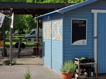 Campingplatz Stadt Koeln Ein blauer Kiosk auf einem Campingplatz mit Schildern für "Garten Café" und "Breakfast Service".A blue kiosk on a campsite with signs for "Garden Café" and "Breakfast Service".