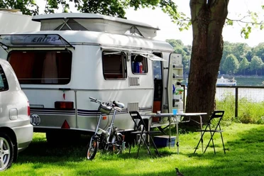 Campsite City of Cologne Camping trailer, table and chairs on a green lawn, with a tree and river in the background.