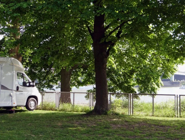Campsite City of Cologne Wohnmobil parkt unter grünen Bäumen, mit Blick auf den Rhein und eine nahegelegene Brücke in Köln.Motorhome parked under green trees, with a view of the Rhine and a nearby bridge in Cologne.