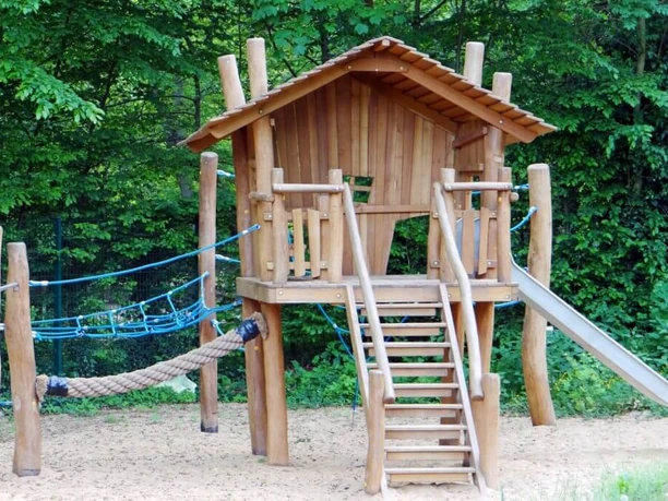 Waldbad und Camping Holzspielhaus mit Rutsche, Seilbrücke und Kletternetz auf Sand umrahmt von üppig grünem Wald.