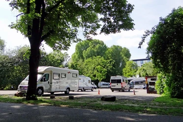 Reisemobilhafen Wohnmobile parken unter schattigen Bäumen, im Hintergrund grünes Laub und strahlend blauer Himmel.