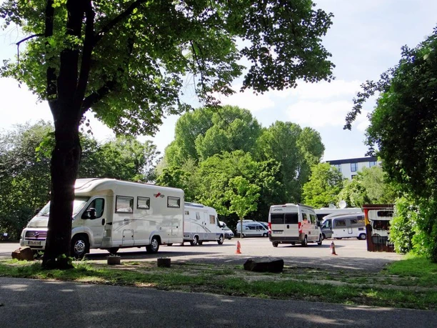 Reisemobilhafen Wohnmobile parken unter schattigen Bäumen, im Hintergrund grünes Laub und strahlend blauer Himmel.