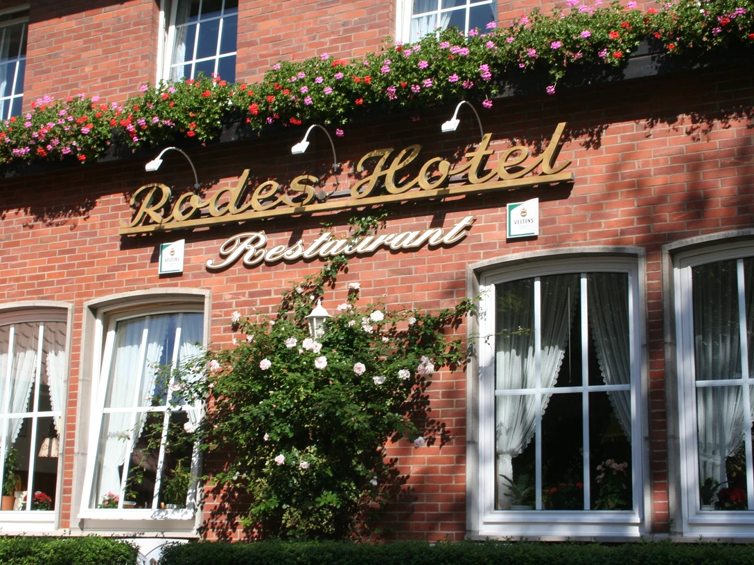 Rodes Hotel Rotes Backsteingebäude mit Blumen geschmückt, darunter der Schriftzug "Rodes Hotel Restaurant".Red brick building decorated with flowers, including the lettering "Rodes Hotel Restaurant".Rød murstensbygning dekoreret med blomster, herunder bogstaverne "Rodes Hotel Restaurant".Gebouw van rode baksteen versierd met bloemen, inclusief het opschrift "Rodes Hotel Restaurant".