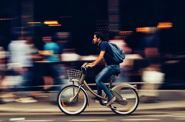 pexels-snapwire-310983(1).jpg Ein Mann fährt in rasanter Geschwindigkeit auf einem silbernen Leihfahrrad, umgeben von verschwommenen, passierenden Fußgängern einer städtischen Straße.