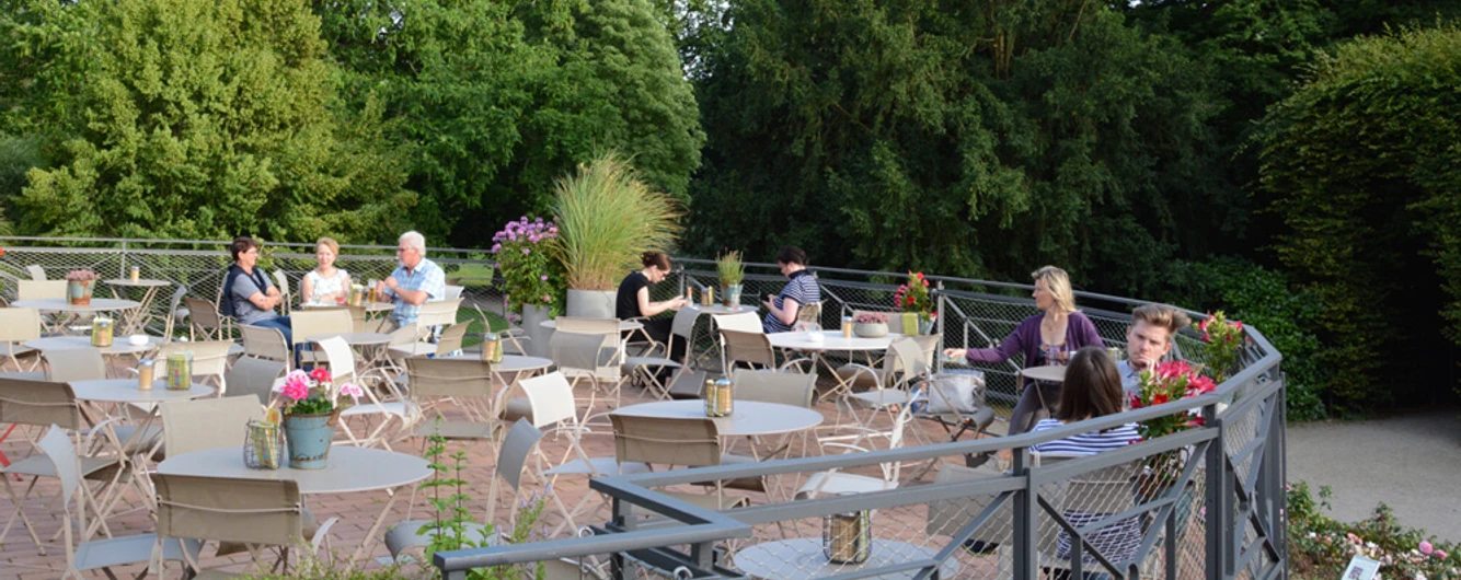 Dank Augusta Außenterrasse mit grüner natürlicher Umgebung. Menschen sitzen an Tische und unterhalten sich.Outdoor terrace with green natural surroundings. People are sitting at tables and talking.