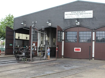 Hallen der Museums-Eisenbahn Bruchhausen-Vilsen Historische Eisenbahnwerkstatt mit geöffneten Toren, Schienen und einer kleinen Gruppe von Menschen.