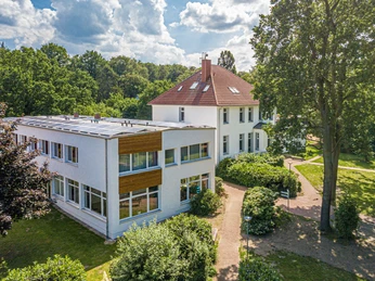 Ev. Heimvolkshochschule Loccum e. V. Ein großes, zweistöckiges Gebäude mit weißen Wänden und rot-braunem Dach, umgeben von grünen Bäumen.