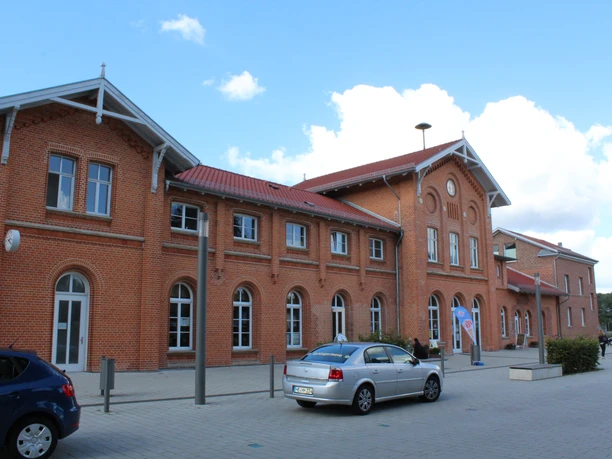 Backstein-Gebäude mit rotem Dach und Rundbogenfenstern, parkende Autos vor blauem Himmel.