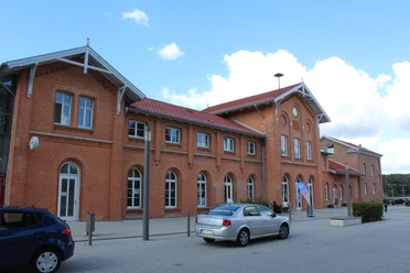 Bahnhof Kirchweyhe Backstein-Gebäude mit rotem Dach und Rundbogenfenstern, parkende Autos vor blauem Himmel.