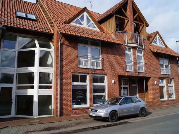 A multi-storey building with a red tiled roof and balconies, a silver car parked on the street in front of it.