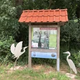 Zwei silberne Reiher-Skulpturen flankieren eine Infotafel mit rotem Dach in einem grünen Waldstück.