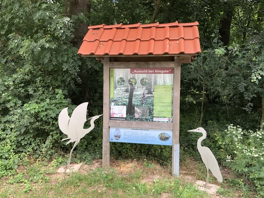 Zwei silberne Reiher-Skulpturen flankieren eine Infotafel mit rotem Dach in einem grünen Waldstück.