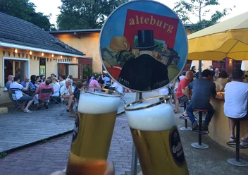Die Alteburg Zwei Kölsch Gläser stoßen an und im Hintergrund sitzen Menschen im Innenhof.