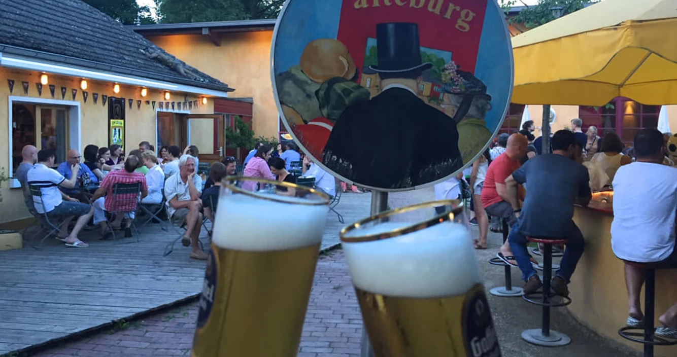 Die Alteburg Zwei Kölsch Gläser stoßen an und im Hintergrund sitzen Menschen im Innenhof.Two Kölsch glasses clink and in the background, people are sitting in the courtyard.