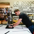 Hülden Ein junger Mann arbeitet konzentriert an einer Industriemaschine, umgeben von Regalen mit Metallteilen.A young man works intently on an industrial machine, surrounded by shelves of metal parts.