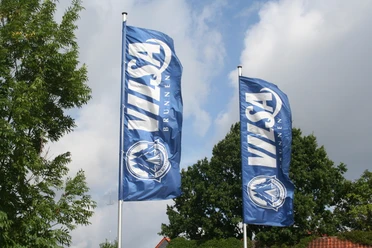 Two blue flags with the "VILSA Brunnen" logo and lettering fly in front of a cloudy sky.