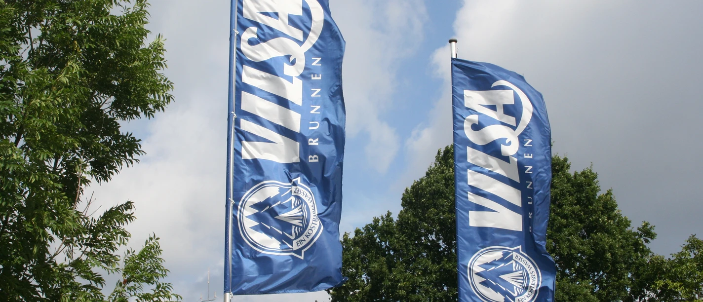 Vilsa Flaggen Zwei blaue Flaggen mit dem Logo und Schriftzug "VILSA Brunnen" wehen vor einem bewölkten Himmel.