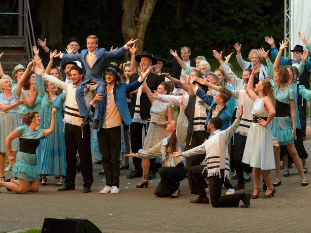 Aufführung auf einer Freilichtbühne mit einer großen Gruppe in farbenfrohen Kostümen und erhobenen Armen.