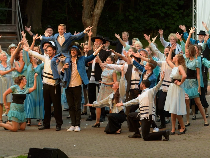 Freilichtbühne in Bökendorf Aufführung auf einer Freilichtbühne mit einer großen Gruppe in farbenfrohen Kostümen und erhobenen Armen.