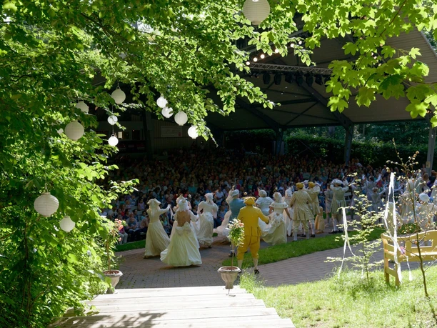 Freilichtbühne Bökendorf: Menschengruppen genießen eine Theateraufführung unter freiem Himmel im Grünen.