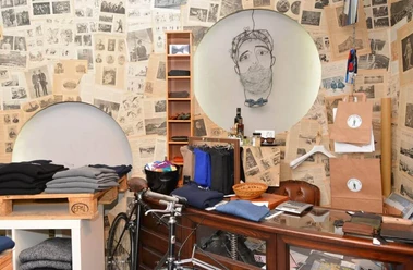 Greenwich Man Time Ein stilvoll eingerichteter Laden mit Vintage-Charme, dekoriert mit alten Zeitungen. Im Fokus stehen elegante Männerkleidung, Accessoires und ein Fahrrad. Ein gezeichnetes Porträt an der Wand. Modern und traditionell vereint.