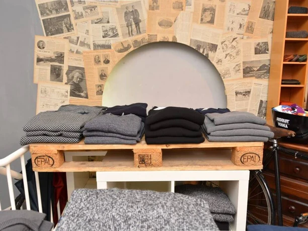 Greenwich Man Time Ein Holzregal aus Paletten, darauf ordentlich gefaltete Pullover in Grautönen und Schwarz, steht vor einer Wand beklebt mit Zeitungsausschnitten.